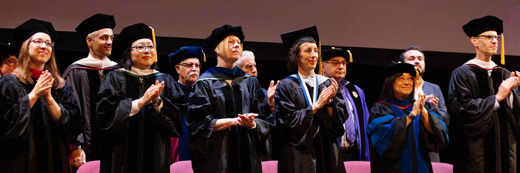 Faculty members dressed in caps and gowns are applauding.